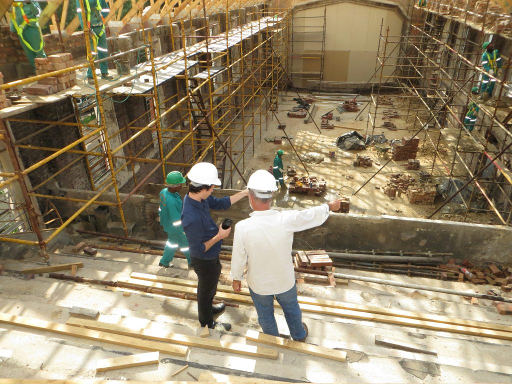 Twee mensen met witte bouwhelmen bespreken de voortgang van een bouwproject, terwijl arbeiders op de achtergrond werken tussen steigers en bakstenen.