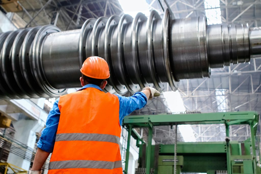 Een fabrieksmedewerker met een oranje veiligheidsvest en helm die een groot metalen onderdeel inspecteert in een industriële werkplaats.